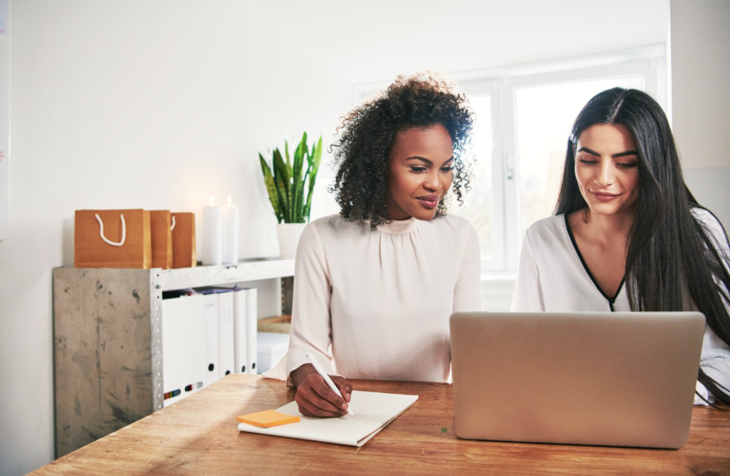 Two women sitting at a laptop and writing notes.