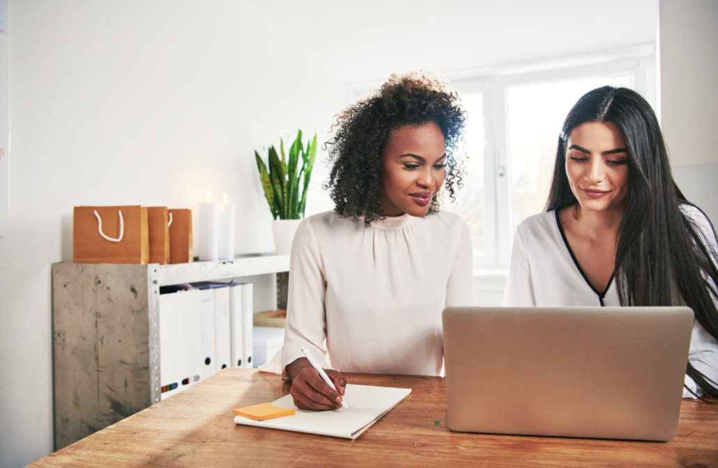 Two women sitting at a laptop and writing notes. 